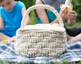 Op de afbeelding: Een crèmekleurige, met de hand gehaakte mand met een geweven patroon en een afgeronde handgreep. De mand staat op een blauw-groen geruite deken. De mand is waarschijnlijk bedoeld voor het dragen van spullen, zoals eten voor een picknick.