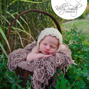 Puede incluir: Un bebé recién nacido con un gorro de crochet blanco está durmiendo en una cesta de mimbre marrón forrada con una manta peluda marrón. La cesta está sentada sobre un lecho de hierba verde.