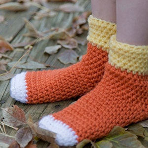 Puede incluir: Un par de calcetines de ganchillo naranja y amarillo con ribetes blancos. Los calcetines están hechos con un punto texturizado y están sobre una superficie de madera con hojas de otoño.
