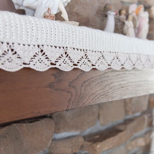 May include: Cream-coloured, crocheted mantel scarf with a scalloped lace edge. The scarf is draped over a dark wooden mantel, with decorative figurines visible in the background. The design features a repeating pattern of openwork triangles.