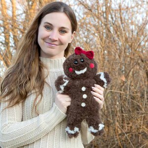 Puede incluir: Una mujer sostiene un juguete de peluche de jengibre marrón con detalles de glaseado blanco y un lazo rojo. El juguete tiene una cara sonriente con ojos negros y mejillas rojas.
