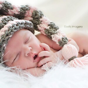 Op de afbeelding: Een slapende pasgeborene met een roze, grijze en witte gehaakte muts met een pompon. De baby ligt op een zacht, wit, pluizig oppervlak. De tekst "Eisells Imagery" is zichtbaar.