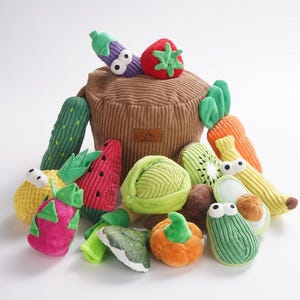 May include: A collection of plush toy vegetables and fruits, including a red strawberry, purple eggplant, green cucumber, and orange carrot. The toys are arranged around a brown corduroy basket, creating a playful and colorful display.