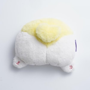 May include: A plush, white and yellow dog butt-shaped pillow. The pillow has a fluffy, soft texture with a yellow top and white bottom. The dog's rear paws are visible, with pink paw pads. A purple tag is on the side.