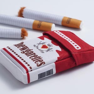 May include: A rectangular red and white fabric pouch designed to resemble a cigarette pack with the text "chewgarettes" printed on the front, accompanied by three fabric cigarette-shaped items with brown fuzzy ends and white bodies.