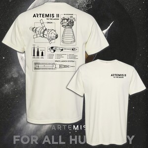 May include: Off-white t-shirts with black graphic designs. One shirt features a detailed diagram of the Artemis II space launch system, including the Orion spacecraft and RS-25 engine. The other shirt has the text "ARTEMIS II TO THE MOON".