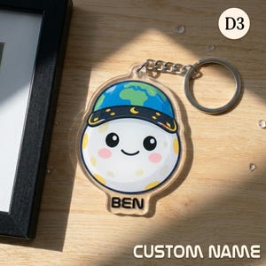 May include: A clear acrylic keychain featuring a cartoon moon character wearing a blue and green earth-themed hat. The keychain has a silver chain and ring. The name "BEN" is printed on the bottom. The words "CUSTOM NAME" are also visible.