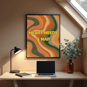 May include: A framed art print with the text "HEART NEEDS A NAP" in yellow letters. The artwork features a retro-style design with wavy stripes in shades of orange, green, and pink. A black desk lamp, a laptop, and a vase with greenery are also visible.