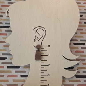May include: A dark brown wooden earring featuring a small circular top piece connected to a larger arch-shaped pendant. The earring is displayed on a wooden profile cutout with a printed ruler showing a length of approximately 1 inch.