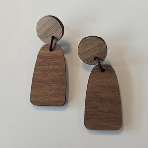 May include: A pair of drop earrings featuring a circular top piece and a larger, elongated arch-shaped bottom piece, both crafted from natural brown wood with visible grain patterns, connected by a thin black metal ring.