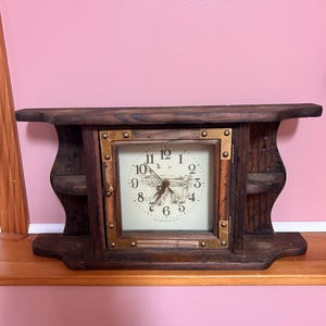 May include: A dark brown wooden mantel clock with a square clock face. The clock face has black numbers and hands, and a small illustration. The clock is framed with brass-colored metal and has a shelf.
