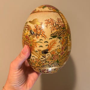May include: An egg-shaped decorative object featuring intricate hand-painted details in gold, red, green, and black. The surface displays a detailed scene with a long-tailed bird, blooming flowers, and foliage, accented with raised gold paint.