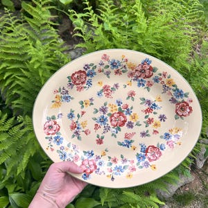 May include: Oval serving platter with a cream-colored background and a floral pattern. The flowers are in shades of red, blue, pink, and yellow. The platter has a green border and is held by a person.
