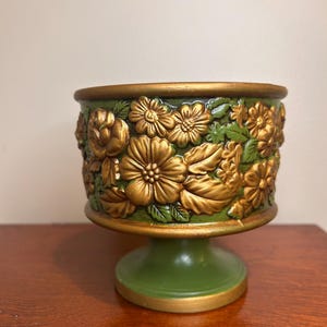 May include: A pedestal planter featuring a deep green base and a cylindrical body adorned with raised, metallic gold floral relief patterns. The rim and the circular foot of the planter are finished with a matching metallic gold trim.