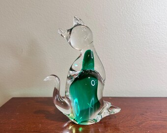 May include: A clear glass cat figurine featuring a smooth, rounded silhouette with a vibrant emerald green core encased within the transparent body. The cat is depicted in a seated position with its tail curled upward and head turned.
