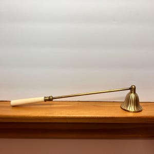 May include: A brass candle snuffer with a bell-shaped head and a long handle. The handle has a light-colored wooden grip. The snuffer is resting on a wooden surface against a white wall.