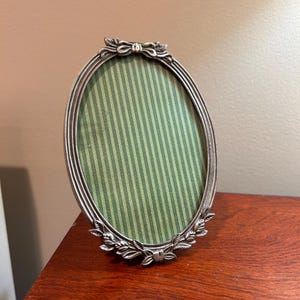 May include: An oval-shaped silver-toned metal picture frame featuring a decorative bow at the top and floral leaf detailing at the bottom. The frame contains a green and dark green vertical striped insert within the oval display area.