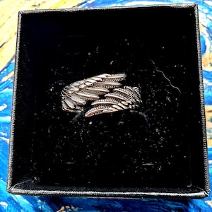 May include: A silver-colored ring shaped like angel wings, displayed in a black box. The ring features detailed feather textures and is presented against a backdrop of blue and yellow brushstrokes. The ring is a jewelry item.