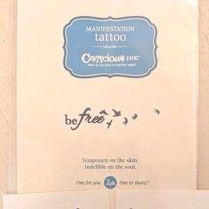 以下が含まれることがあります： 一時的なマニフェストタトゥーのパッケージ。パッケージには、「MANIFESTATION tattoo」と「Conscious INK」の文字が青と白のデザインで表示されています。「be free」というフレーズは筆記体で書かれており、周りを鳥が飛んでいます。「Temporary on the skin. Indelible on the soul.」というテキストも含まれています。