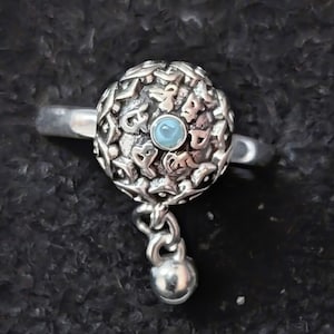 May include: A silver ring featuring a detailed, patterned sphere with a small, light blue gemstone at its center. A small silver ball hangs from the sphere by a chain. The ring is set against a dark, textured background.