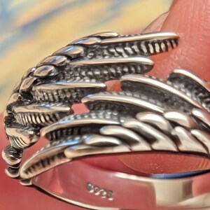 May include: Close-up of a silver ring with a detailed, textured design resembling a wing or feather. The ring has a polished finish and a band with the marking "925". The ring is likely made of sterling silver.