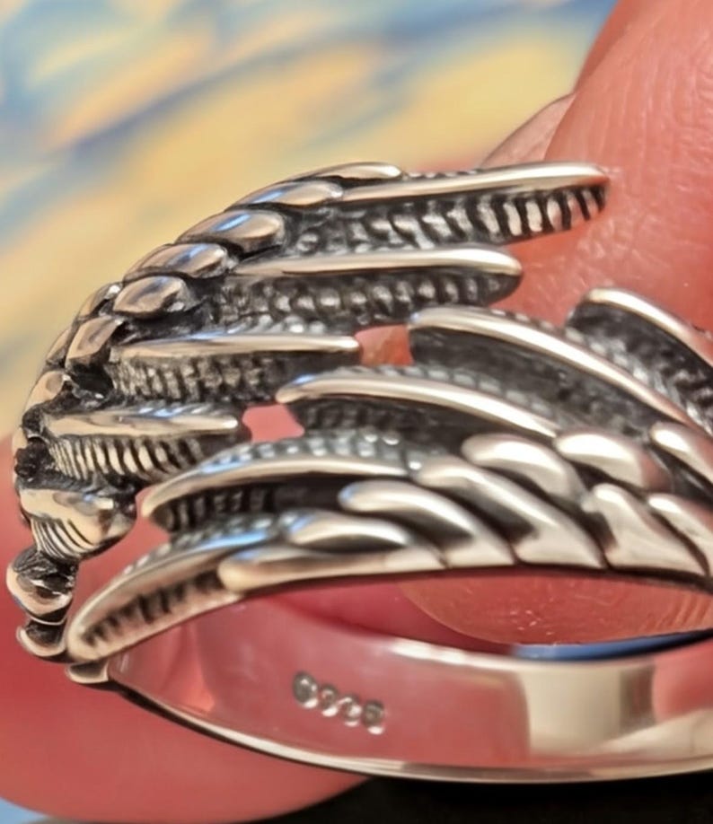 May include: Close-up of a silver ring with a detailed, textured design resembling a wing or feather. The ring has a polished finish and a band with the marking "925". The ring is likely made of sterling silver.