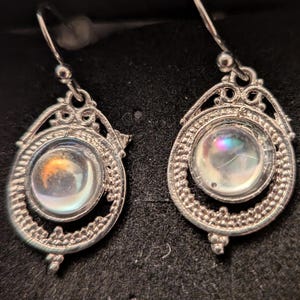 May include: A pair of silver-toned earrings with a teardrop design. Each earring features a round, iridescent stone set within an ornate, openwork frame. The frames have a detailed, textured border and decorative elements.