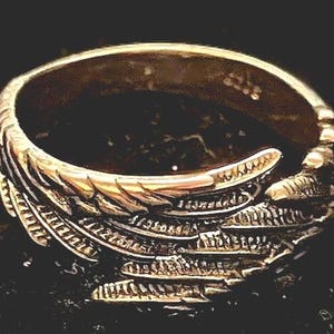 May include: A gold-colored ring with a detailed wing design. The ring features intricate feather detailing, creating a textured appearance. The ring is set against a dark background, highlighting its metallic sheen. A piece of jewelry.