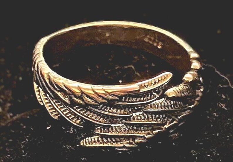 May include: A gold-colored ring with a detailed wing design. The ring features intricate feather detailing, creating a textured appearance. The ring is set against a dark background, highlighting its metallic sheen. A piece of jewelry.