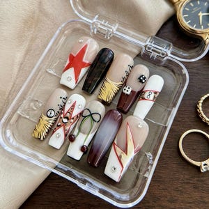 May include: A clear plastic case holds a set of ten long, decorated fingernails. Designs include a red star, black and gold patterns, dice, and a bow. A gold watch and two rings are also visible.
