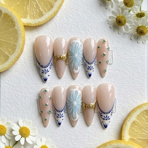 Puede incluir: Un conjunto de uñas postizas en forma de almendra con una base rosa claro. Las uñas presentan varios diseños, incluyendo patrones florales azules y blancos, detalles dorados y adornos de piedras preciosas verdes. Las uñas están dispuestas sobre una superficie blanca con rodajas de limón y margaritas.