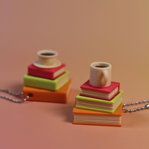 May include: Two pendant necklaces, each featuring a stack of colorful miniature books topped with a small coffee cup. The books are orange, green, and red. The coffee cups are white with dark brown coffee inside. The background is a gradient of pink and orange.