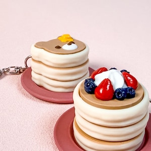 May include: Two pancake-shaped keychains. One keychain features a bear-shaped pancake with a yellow pat of butter. The other keychain has pancakes topped with whipped cream, blueberries, and strawberries. Both keychains are on pink plates.