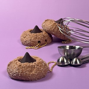 May include: Three brown, donut-shaped keychains with a dark chocolate kiss in the center, and a gold chain. A stainless steel whisk, measuring spoons, and a small bowl are on a purple background.