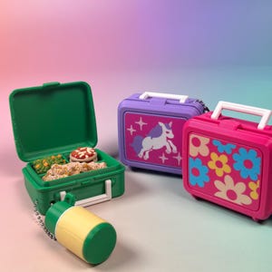 May include: Three miniature lunchbox keychains with white handles. One green box is open, revealing tiny food items like pizza and vegetables, with a yellow and green thermos attached. A purple box features a white unicorn, and a pink box has floral patterns.