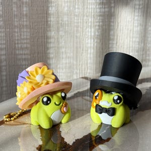 Könnte beinhalten: Zwei skurrile Froschfiguren. Ein Frosch trägt einen Hut mit gelben Blumen und einem rosa Rand. Der andere Frosch ist mit einem schwarzen Zylinder und einer schwarzen Fliege bekleidet. Beide Frösche sind lindgrün mit weißem Bauch.