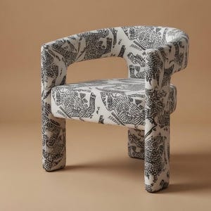 Könnte beinhalten: Kleiner, gepolsterter Sessel mit abgerundeter Rückenlehne und Armlehnen. Der Stuhl ist mit einem weißen Stoff mit einem schwarzen, abstrakten Muster bezogen. Der Stuhl hat vier kurze Beine und steht vor einem neutralen Hintergrund.