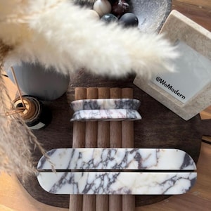 Op de afbeelding: Een decoratieve opstelling met marmeren en houten elementen. Een wit en grijs marmeren dienblad ligt op houten deuvels, met een bijpassend marmeren stuk erboven. Een kom met decoratieve stenen bollen en pampasgras maken het geheel compleet.