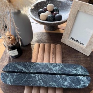 May include: A still-life arrangement featuring home decor items. A dark green marble tray is in the foreground, with a wooden tray and a stone bowl filled with decorative spheres. A framed photo and a gray vase are also present.