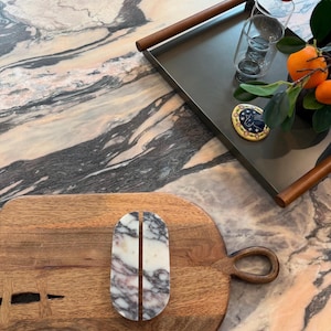 May include: A wooden cutting board with a handle and a pair of marble coasters. The coasters are white with gray and purple veining. A dark gray tray with wooden handles holds glasses and oranges.
