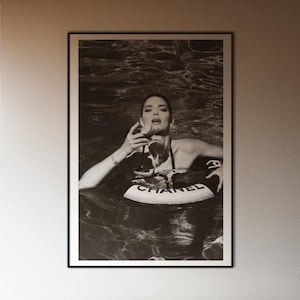 May include: A black and white framed print of a woman in a pool with a Chanel floatie hangs above a bed with a dark gray comforter. The room has large windows and a wooden floor.