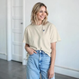 Puede incluir: Camiseta color crema con un pequeño diseño bordado azul en el pecho. La camiseta tiene mangas cortas y se combina con vaqueros de mezclilla azul claro. La modelo sonríe y lleva una pulsera de plata.