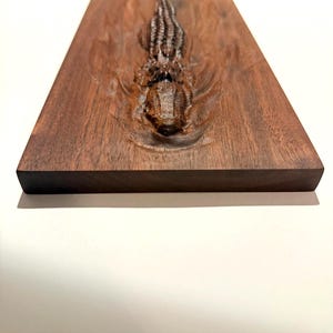May include: A rectangular, dark brown wooden plaque with a carved alligator design. The alligator is depicted in a detailed, three-dimensional relief, with textured scales and open jaws. The wood grain is visible, adding to the natural aesthetic.