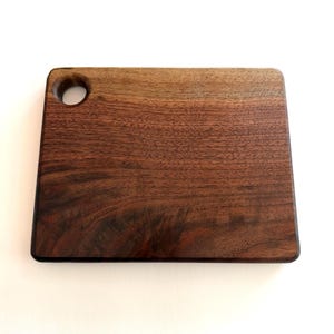 May include: A rectangular wooden cutting board with a dark brown finish. The board has rounded corners and a circular hole in the upper left corner. The wood grain is visible throughout the board.