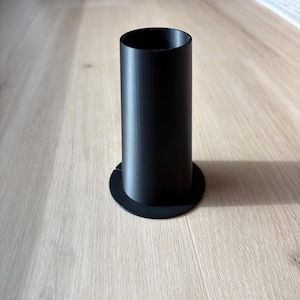 May include: A black cylindrical object with a circular base. The object is made of a solid material and stands upright on a light-colored wooden surface. The cylinder has a smooth, matte finish.