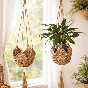 May include: Two hanging plant holders made of woven natural fiber. Each holder is suspended from a wooden rod by a rope and metal hook. One holder contains a green plant in a glass jar. The other is empty. Both have a decorative tassel.