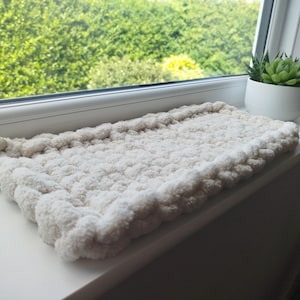 May include: A rectangular, off-white chunky knit mat featuring a thick, soft, looped yarn texture. The mat has a small, light brown leather-like tag attached to one side, showcasing a simple, hand-crafted design with a raised, plush surface.