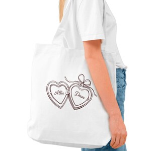 May include: White tote bag featuring a brown heart-shaped locket design with the names "Allie" and "Dean" inside. The locket is connected with a bow. The bag is carried by a person wearing a white t-shirt and blue jeans.