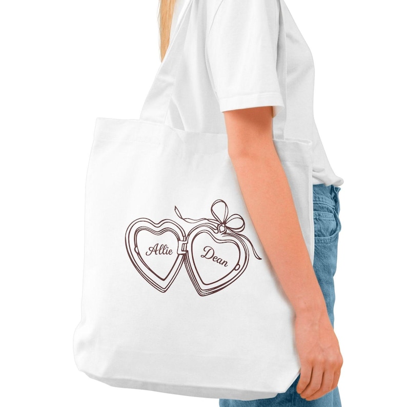 May include: White tote bag featuring a brown heart-shaped locket design with the names "Allie" and "Dean" inside. The locket is connected with a bow. The bag is carried by a person wearing a white t-shirt and blue jeans.
