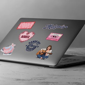 May include: A laptop covered in stickers. Stickers include a pink cassette tape with the word "Crushing", a pink heart with the text "Garrett Graham", and a sticker of a woman playing a guitar. The laptop is on a gray surface.